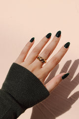 A hand wearing a gold croissant ring with a unique twisted design, styled against a soft beige background with dark green nails, and a cozy green sweater.