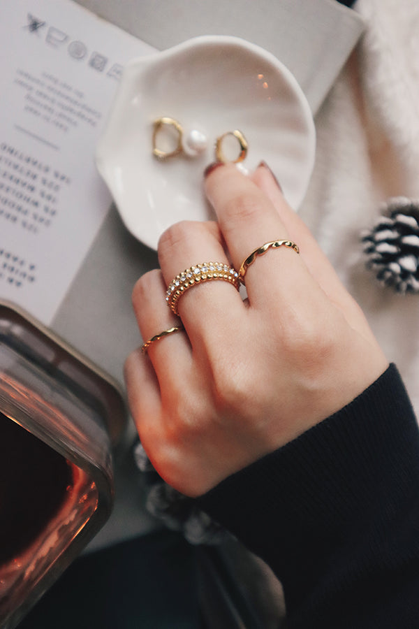 A hand adorned with three elegant gold rings is placed next to a dish holding earrings, creating a cozy, refined look.