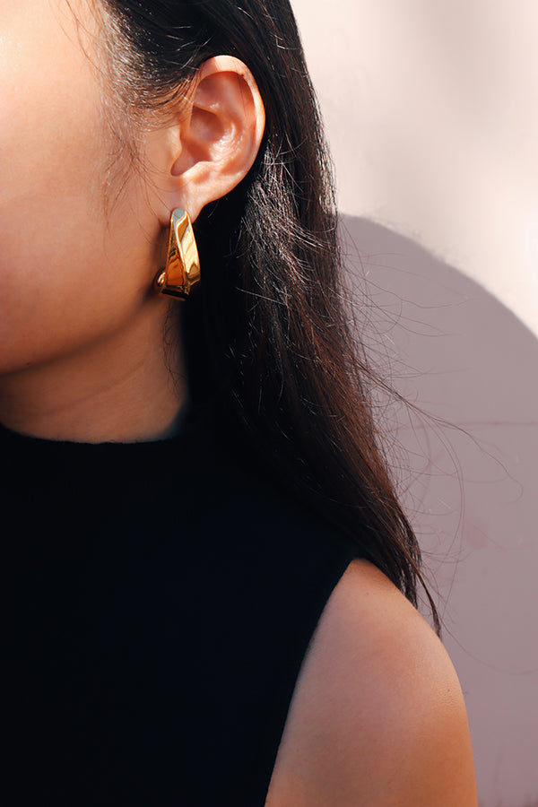 A close-up of a woman wearing bold, sculptural gold earrings paired with a black sleeveless top, showcasing the modern and sleek design of the jewelry.