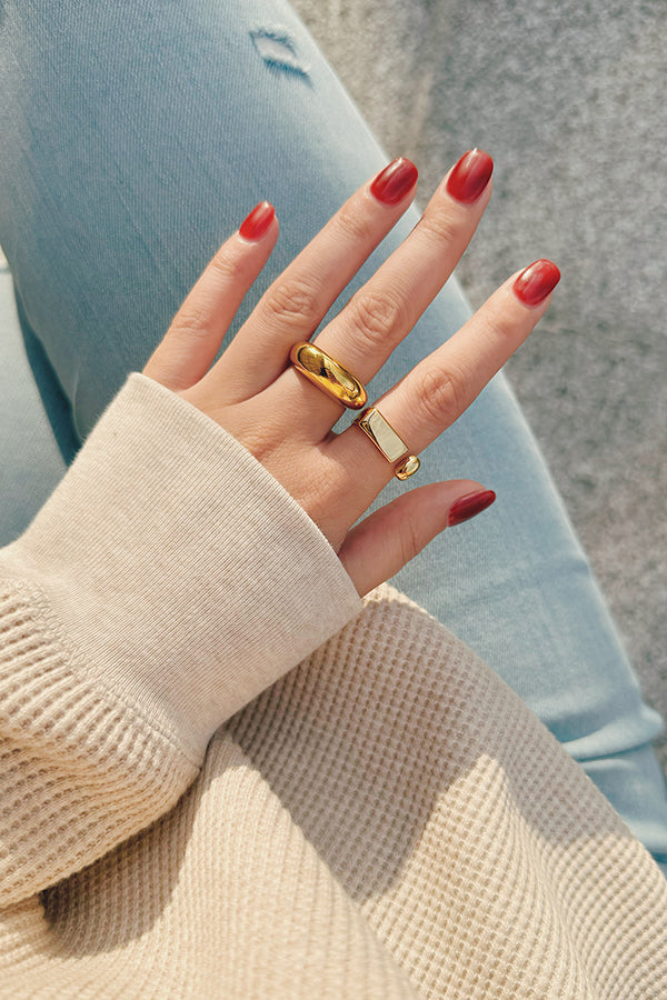 A close-up of a hand with red nails, wearing two chunky gold statement rings, styled with a beige ribbed sweater and light blue jeans.