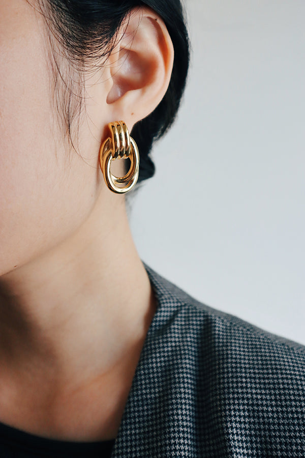 Close-up of a woman wearing chunky gold geometric earrings paired with a gray checkered blazer, highlighting a modern, bold accessory style.