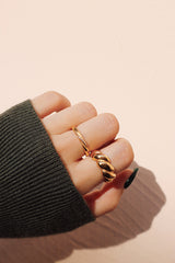 Close-up of a hand wearing a chunky twist ring and a delicate gold band, against a warm-toned background. The fingers are partially wrapped in a green knit sleeve.