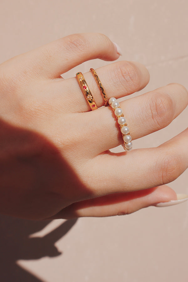 A hand wearing three elegant rings: one pearl band, a sleek twisted gold ring, and a gold ring with subtle star and gemstone detailing, creating a delicate and chic stacked look.