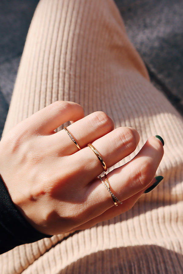 A hand adorned with three slim, minimalist gold stacking rings, catching natural light and creating a sophisticated look against a beige ribbed fabric background.