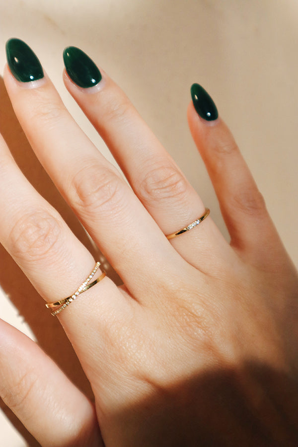 Close-up of a hand wearing two thin gold stacking rings with subtle diamond accents, highlighting elegant green nail polish and soft lighting on the skin.