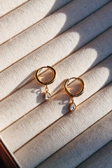 A pair of gold hoop earrings with delicate teardrop crystal pendants displayed on a soft, textured surface, casting gentle shadows in natural sunlight.