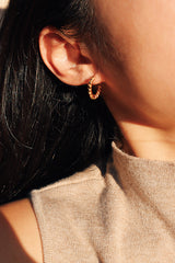 A woman wearing a twisted gold hoop earring, accentuating her warm beige top with soft natural lighting.