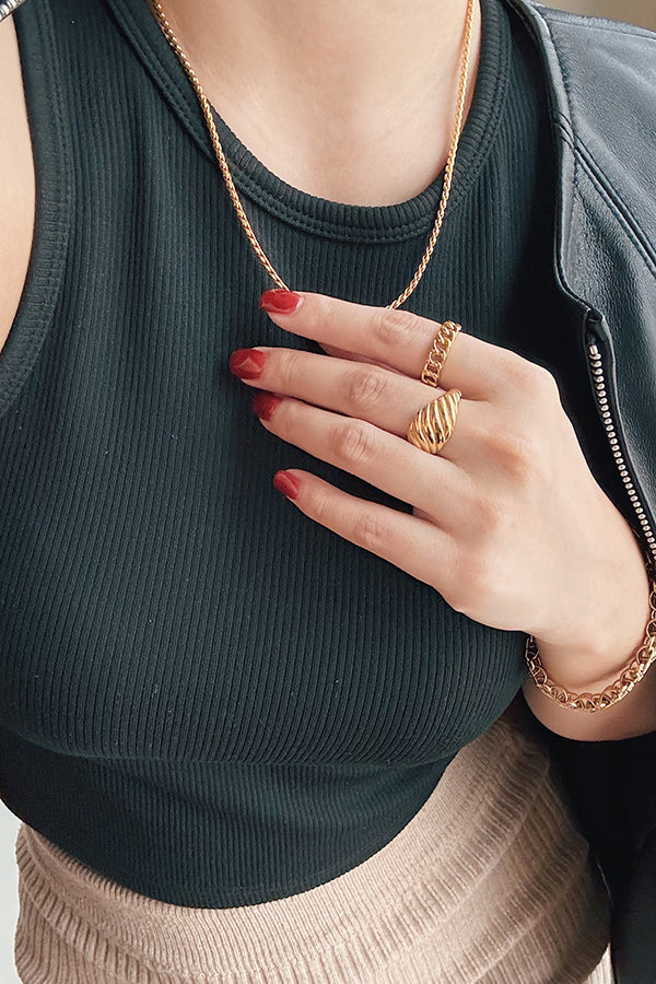 A close-up of a woman wearing a gold chain necklace, gold textured ring, and gold link bracelet, styled with a black ribbed top and leather jacket for a modern and chic look.