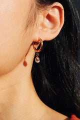 Close-up of a gold hoop earring featuring a delicate teardrop cz dangle, worn on a woman's ear against a softly lit background.