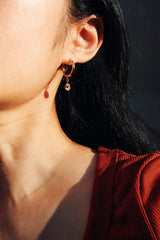 A woman wearing a gold hoop earring with a dangling teardrop-shaped cubic zirconia, catching warm sunlight against a dark background and a red top.