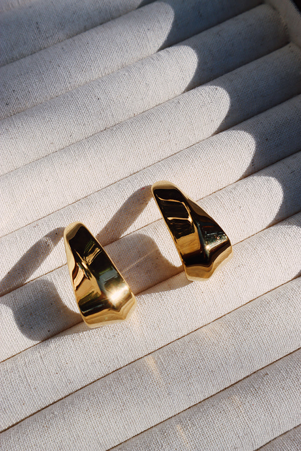 A pair of modern gold hook earrings with a sleek, curved design placed on a neutral beige fabric display, reflecting natural sunlight.