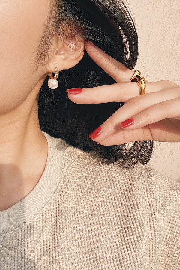 Close-up of a woman's ear wearing a pearl stud earring with a gold hoop accented by small stones, alongside her hand showcasing minimalist gold rings with a glossy red manicure.