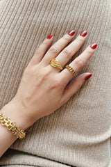 A hand adorned with a bold gold statement ring and chain link ring, paired with a chunky gold bracelet against a textured beige background.