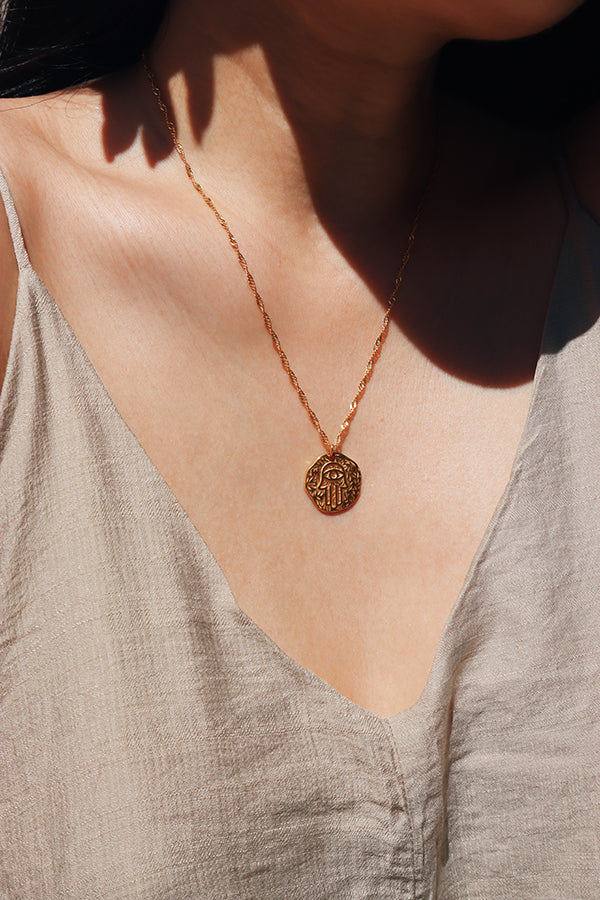 A close-up view of a womans neckline adorned with a delicate 18K gold-plated necklace featuring a hand of Fatima pendant, paired with a light beige linen dress.