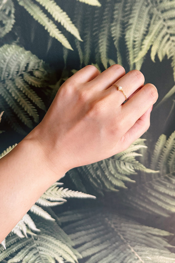 A close-up image of a hand wearing a delicate gold chain ring featuring a small pearl, set against a background of lush green fern leaves.