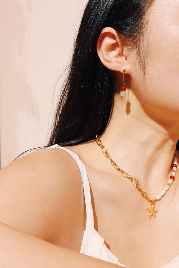 Side view of a woman wearing a dangle earring featuring red agate and glass beads, paired with a chunky gold chain necklace adorned with a starfish charm.