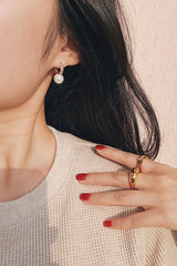 Woman wearing pearl drop hoop earrings and a chunky gold ring, styled with red nail polish and a cream textured top.