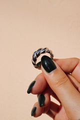 Close-up of a hand holding a silver croissant-shaped ring, showcasing its braided texture against a beige background.