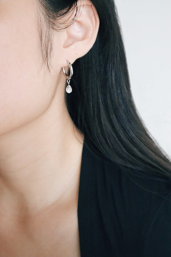 Side profile of a woman wearing a silver hoop earring with a delicate, teardrop-shaped stone dangling from the hoop, complementing her sleek, black attire.