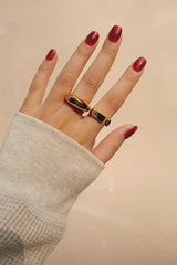 Hand adorned with two statement gold rings featuring black accents, against a neutral background.