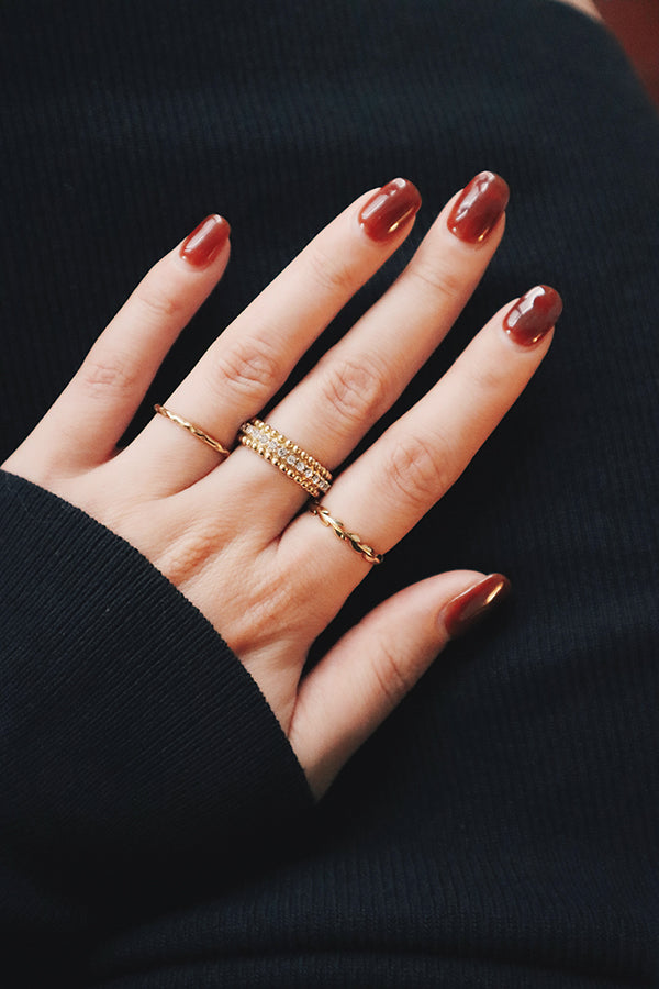 A hand with three elegant gold rings, featuring various textures and designs, resting against a dark background with polished red nails.