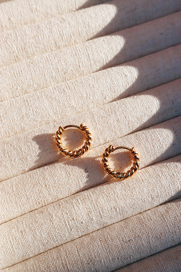 Pair of twisted gold hoop earrings placed on a textured fabric surface, with soft sunlight casting shadows.