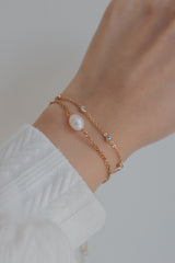 Close-up of a wrist adorned with a delicate 18K gold-plated bracelet featuring evenly spaced cubic zirconia stones, paired with a gold chain bracelet with a pearl centerpiece, worn with a white textured sleeve.