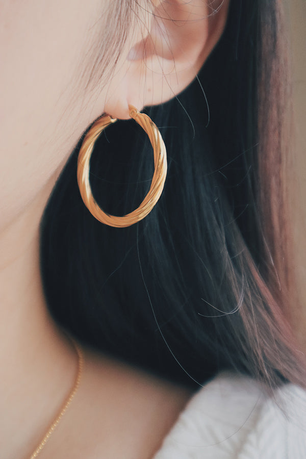 A detailed image of a woman wearing large, 18K gold-plated hoop earrings with a twisted texture design. The earrings catch the light beautifully, highlighting their luxurious finish and textured surface.