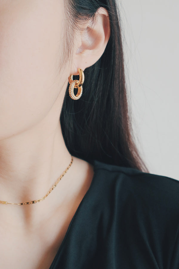 A close-up image of a woman wearing gold hoop earrings with a dangling circular charm. The charm is outlined with cubic zirconia stones, adding a glittering accent to the bold, gold hoop design, perfect for both casual and formal occasions.