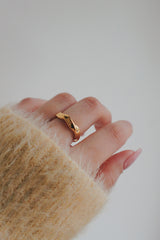 A close-up image of a womans hand wearing a gold wave ring. The ring has a sleek, wave-like design that wraps elegantly around the finger, with a shiny gold finish that complements her outfit.