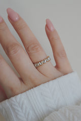 A hand with a light pink manicure is adorned with a gold eternity ring featuring baguette-cut cubic zirconia stones. The rings design showcases a series of rectangular stones set side by side, creating a continuous band of sparkle. The soft knit sleeve in the background adds a cozy touch, contrasting beautifully with the ring's luxurious and shiny appearance.