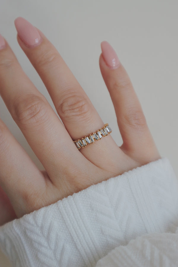 A hand with a light pink manicure is adorned with a gold eternity ring featuring baguette-cut cubic zirconia stones. The rings design showcases a series of rectangular stones set side by side, creating a continuous band of sparkle. The soft knit sleeve in the background adds a cozy touch, contrasting beautifully with the ring's luxurious and shiny appearance.