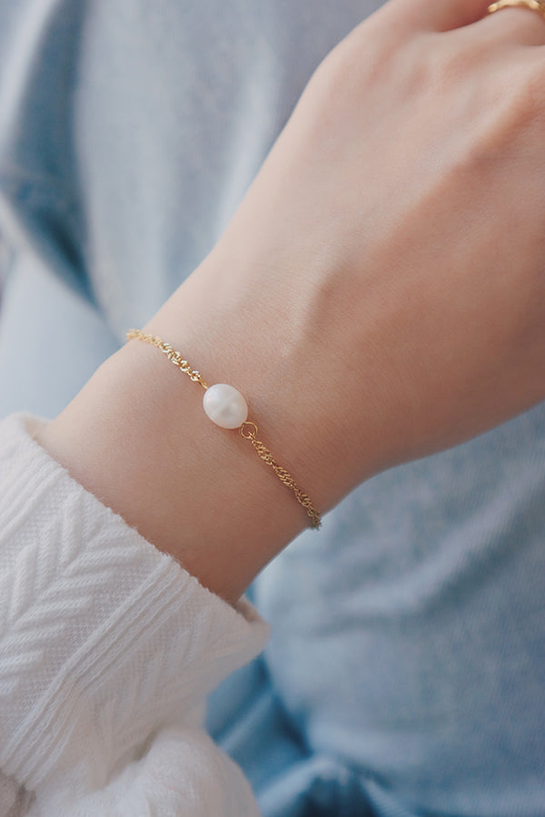 Close-up of a wrist adorned with an 18K gold-plated bracelet featuring a freshwater pearl, styled with a white sweater and gold rings, showcasing a chic and elegant look.