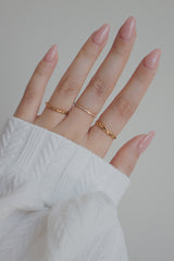 A hand adorned with three gold rings, each with a unique design, showcasing a minimalist and chic style. The middle finger features a slim gold ring with a row of sparkling cubic zirconia stones, while the other fingers sport twisted and plain gold bands. The soft lighting and neatly manicured nails complement the rings, elegant and modern aesthetic.