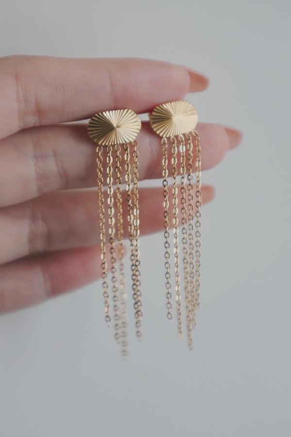 A close-up image of a womans hand holding a pair of fashionable tassel earrings. The earrings feature a sunburst design on the stud with multiple delicate gold chains cascading down, creating a chic and modern accessory.