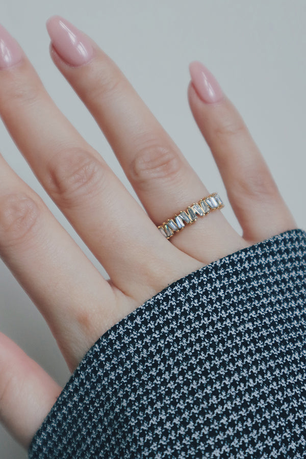 A close-up of a hand wearing a gold eternity ring with baguette-cut cubic zirconia stones. The rings wide band and brilliant stones reflect light elegantly, making it a bold and glamorous accessory. The hand's houndstooth-patterned sleeve adds a stylish and modern contrast to the ring's timeless design.