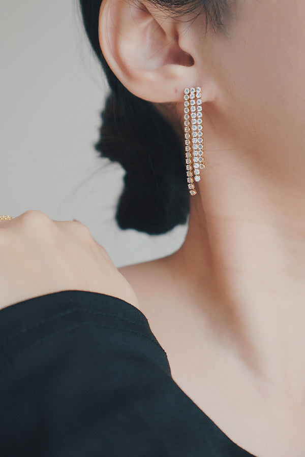 A close-up shot of a womans ear adorned with luxurious tassel earrings made of 18K gold and cubic zirconia stones. The earrings have a sleek, elongated design with multiple rows of sparkling stones, perfect for adding a statement to evening wear.