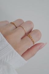 A side view of a hand wearing a trio of gold rings, focusing on the slim band with cubic zirconia stones on the middle finger. The intricate design of the rings and the clean, minimalist look of the nails and white sweater create a harmonious and stylish composition, perfect for showcasing the delicate and refined jewelry.