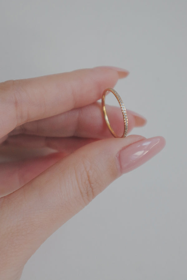  A close-up image of a hand holding a delicate gold ring adorned with tiny cubic zirconia stones. The ring's slim band is highlighted by the sparkling stones, creating a subtle yet elegant look. The hand's neatly manicured nails and soft lighting enhance the ring's refined appearance, making it an ideal accessory for a minimalist and sophisticated style.