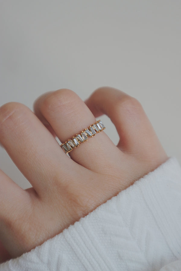 A detailed view of a hand wearing a chunky gold eternity ring encrusted with baguette-cut cubic zirconia stones. The rings thick band and sparkling stones make a statement, while the soft knit sleeve enhances the ring's luxurious appeal, adding a touch of sophistication to the overall look.