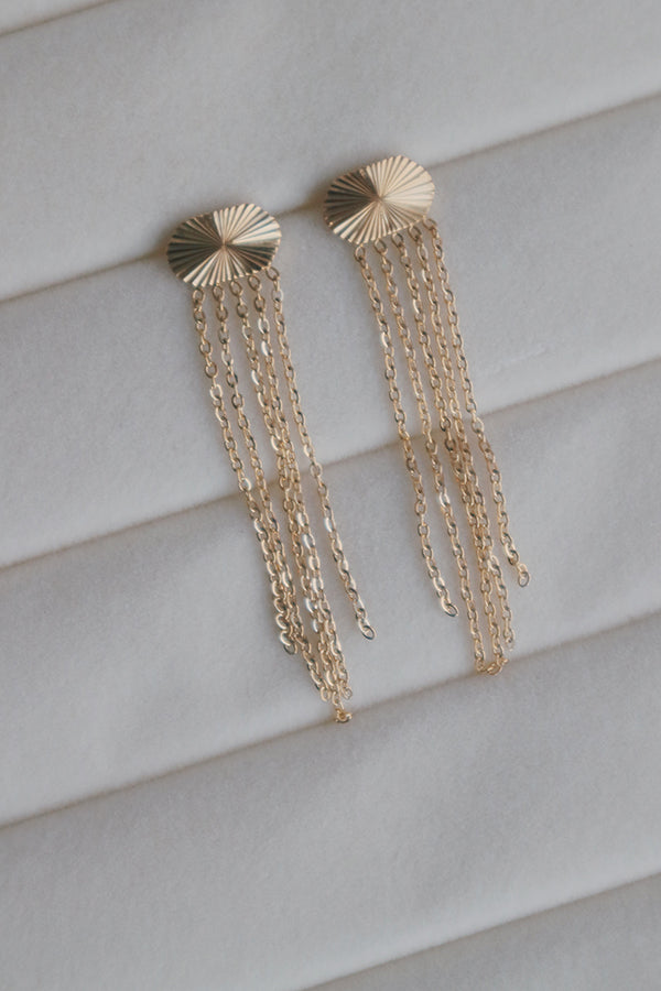 A detailed image of gold tassel earrings laid flat. The earrings showcase a unique sunburst design on the stud, with long gold chains that create a dramatic and glamorous effect.