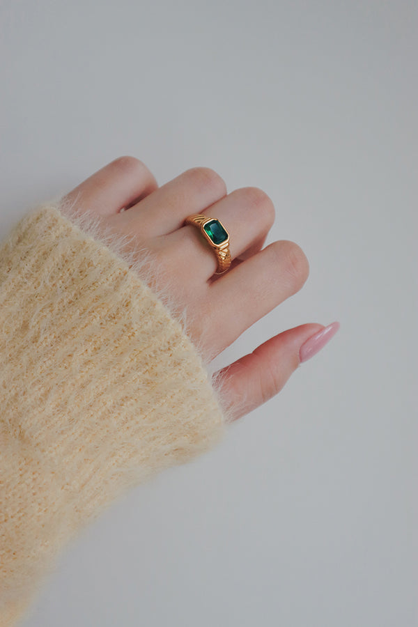 A close-up image of a gold ring with a green cubic zirconia stone. The rings luxurious design, with a richly colored stone set in a detailed band, adds a touch of elegance and sophistication to any outfit.