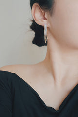 A product image of gold tassel earrings, displayed flat to highlight their cascading design. The earrings feature rows of cubic zirconia stones set in a gold framework, creating a luxurious and eye-catching accessory for evening events.