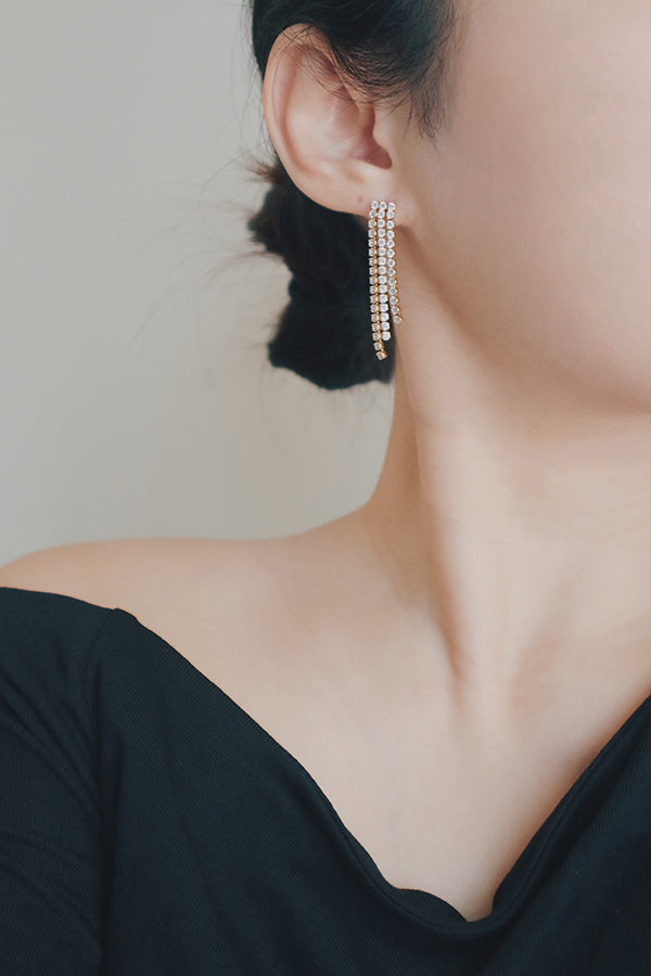 A product image of gold tassel earrings, displayed flat to highlight their cascading design. The earrings feature rows of cubic zirconia stones set in a gold framework, creating a luxurious and eye-catching accessory for evening events.