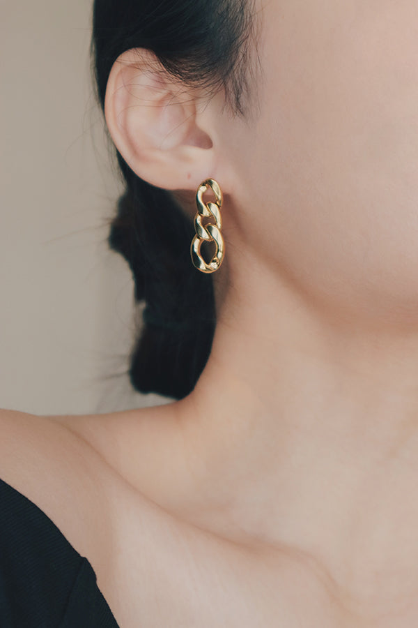 A detailed photo showing a pair of Cuban chain gold earrings in a womans hand. The earrings are designed with chunky, polished gold links, arranged in a chain-like pattern, giving them a bold and contemporary style. The shiny finish adds a sleek and modern touch to the overall design.