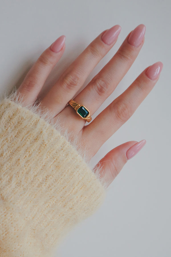 A detailed shot of a gold-plated ring featuring a rectangular green cubic zirconia stone. The rings intricate band and vibrant stone create a striking look, making it an ideal piece for stacking or wearing as a bold statement.