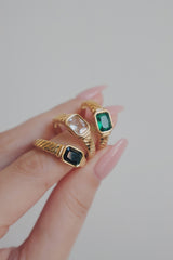 A close-up image of three gold-plated rings held between the fingers, each featuring a different colored cubic zirconia stone: one clear, one green, and one black. The rings have a vintage-inspired design with a rectangular stone set in a detailed band, offering versatile options for stacking or wearing alone.