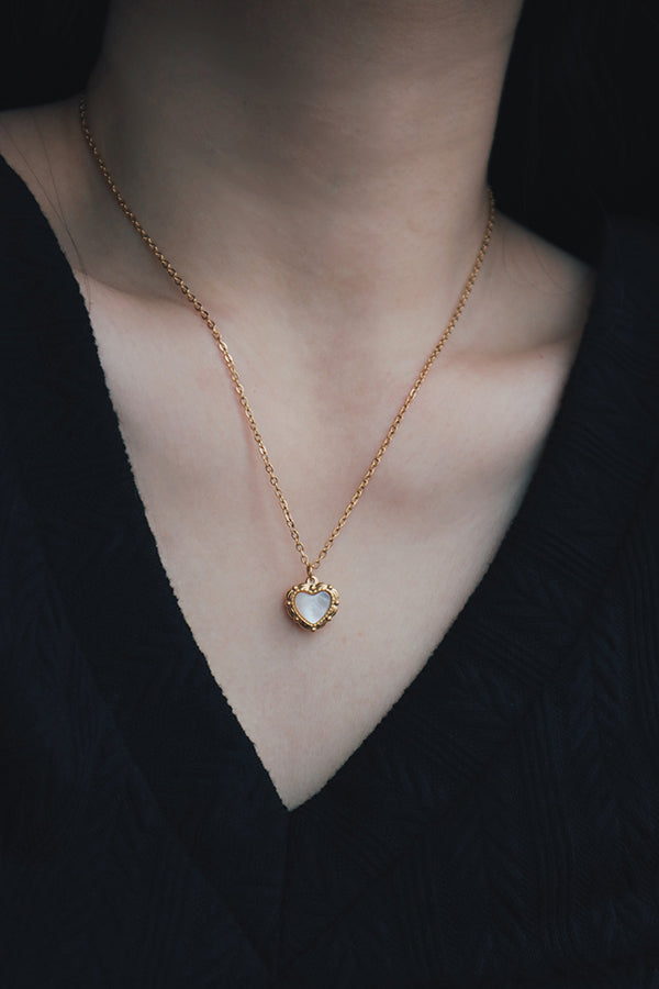 Close-up view of a single gold necklace featuring a heart-shaped pendant with a mother-of-pearl inlay, worn with a black textured V-neck top.