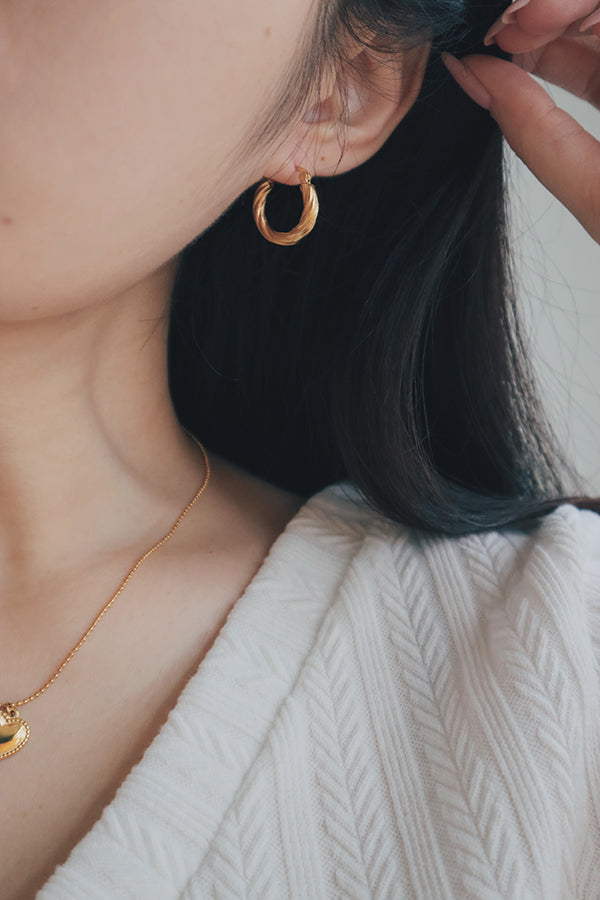 A close-up photo of a woman's ear adorned with a small, twisted texture gold hoop earring. The earring's intricate design and hypoallergenic material make it both stylish and suitable for sensitive ears.