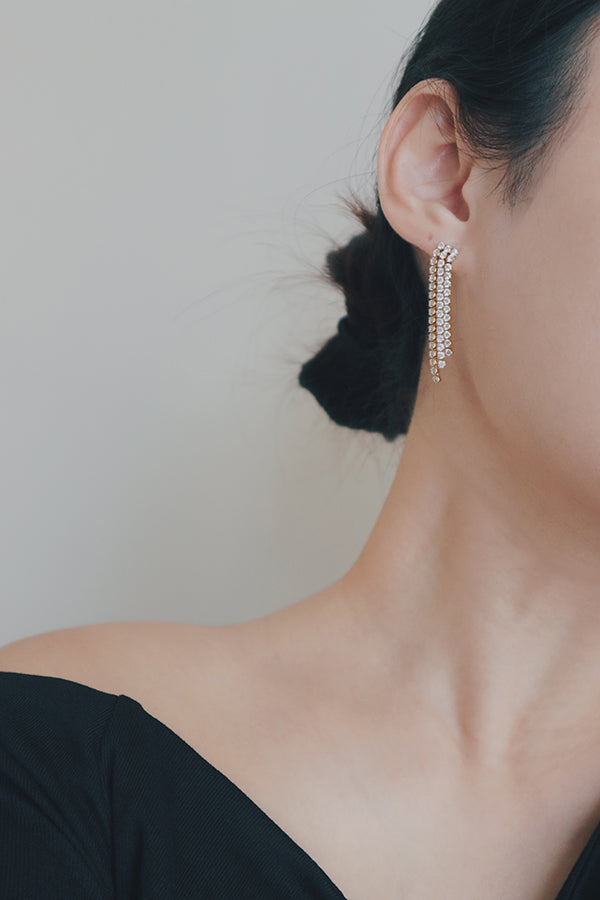 A close-up image of a womans ear wearing a pair of long, cascading tassel earrings. The earrings feature multiple rows of cubic zirconia stones set in a gold base, creating a shimmering, elegant effect. These hypoallergenic earrings add a touch of glamour and sophistication to any outfit.